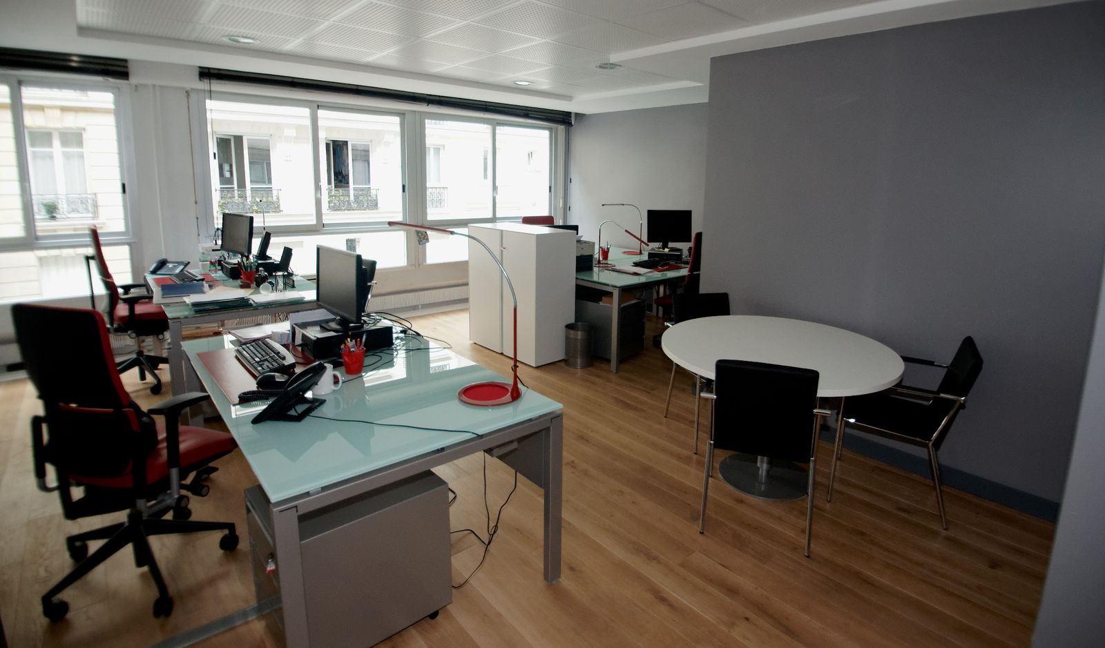 Office floor: glass desks, red chairs, round meeting table, large windows.