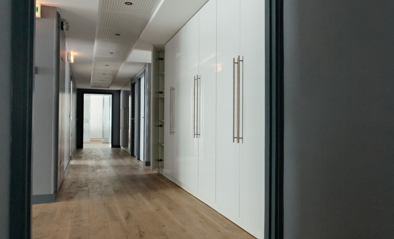 Corridor lined with full-height white lacquered storage, perforated acoustic ceiling, parquet floor.