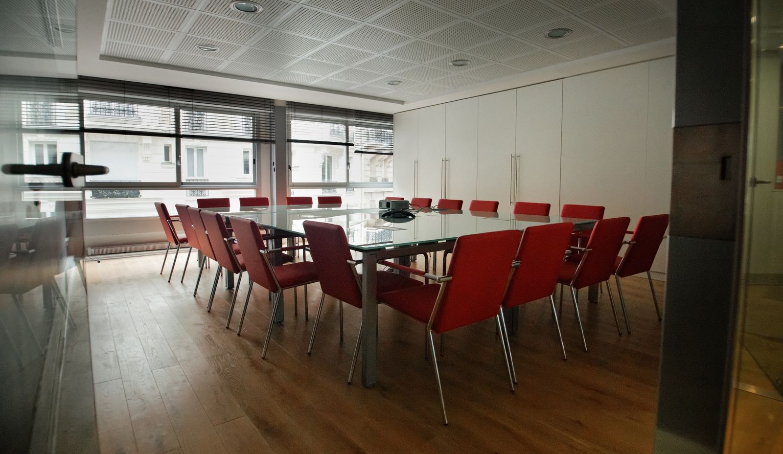 Meeting room: large glass table, around twenty red chairs, windows with blinds overlooking Parisian façades, full-height white storage.
